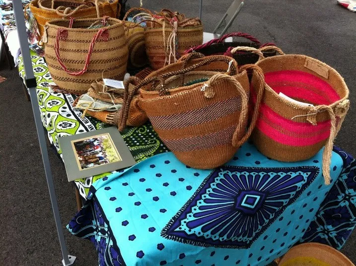 Woven baskets with various patterns and colors are displayed on a table covered with vibrant, patterned fabrics at an outdoor market. A framed photo sits among the baskets.