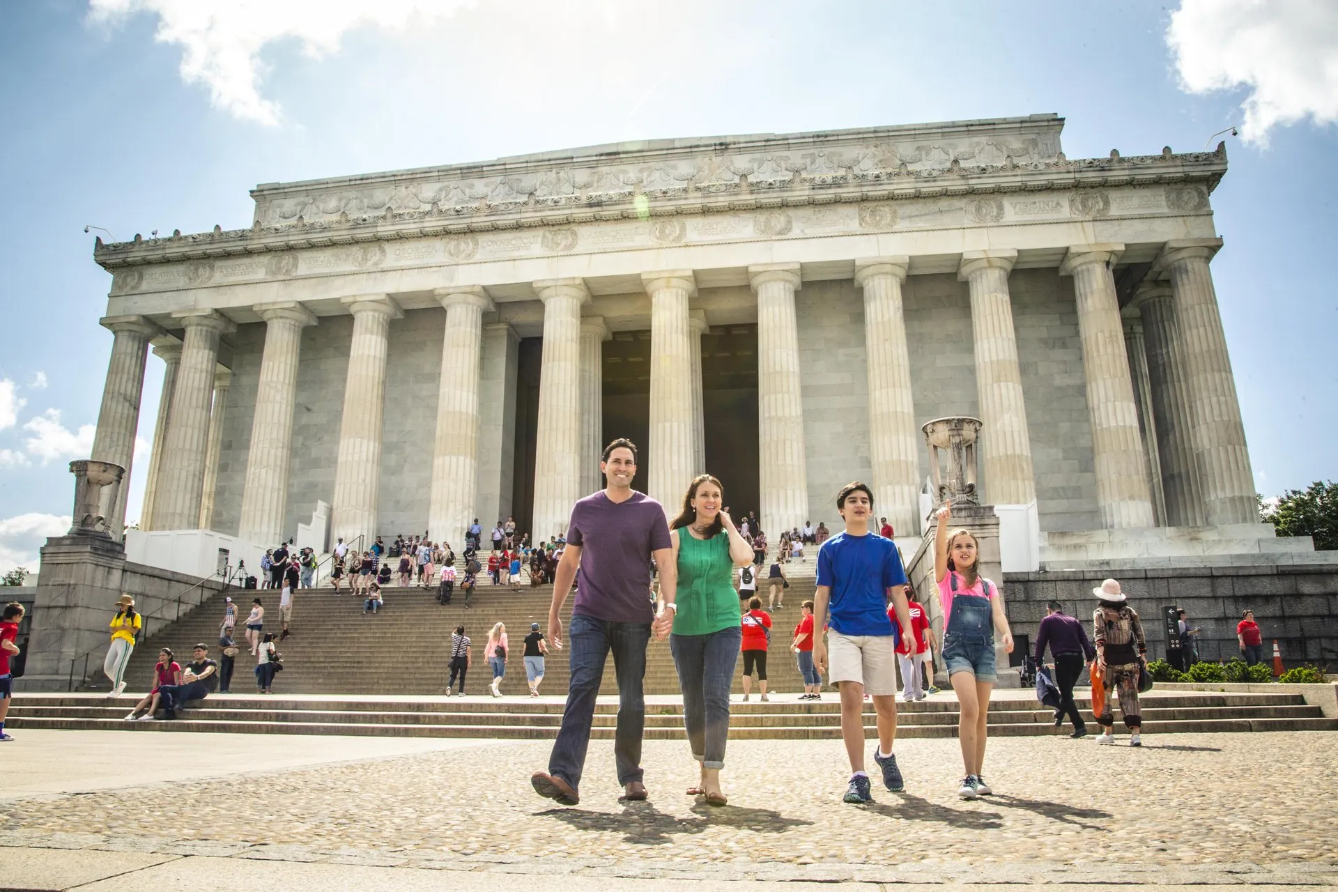 A family of four walks together in front of the Lincoln Memorial, with people sitting and standing on the steps, on a sunny day.