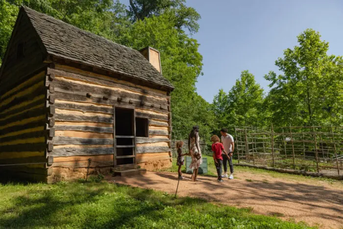 A family of four stands outside a small historic log cabin surrounded by greenery and trees on a sunny day. A wooden garden structure is visible in the background.
