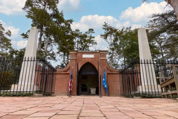 A brick mausoleum with a wrought iron gate, flanked by two flagpoles with American and blue flags, is surrounded by trees under a partly cloudy sky.
