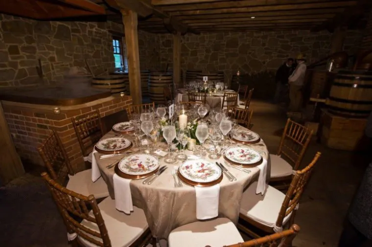A round table set for a formal dinner with floral-patterned plates, wine glasses, and a candle centerpiece in a rustic room with stone walls, wooden beams, and brown chairs.