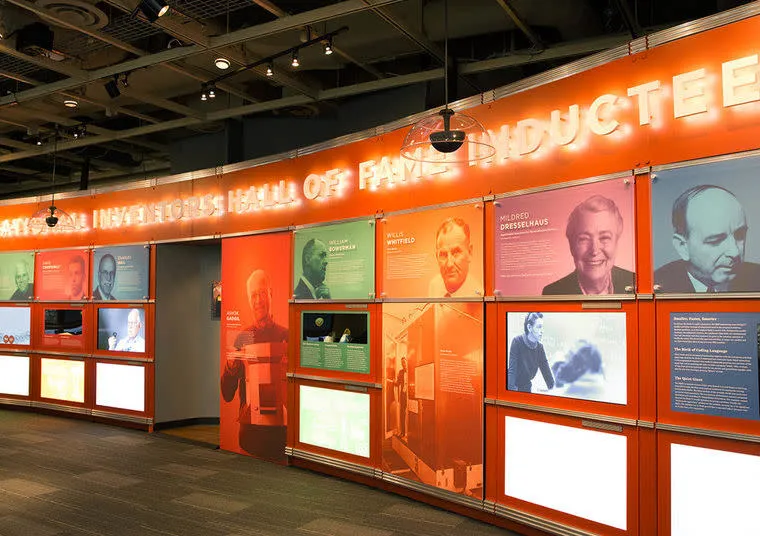 A brightly lit museum exhibit wall showcases the National Inventors Hall of Fame inductees, featuring portraits, names, and achievements of famous inventors with digital displays and text panels.