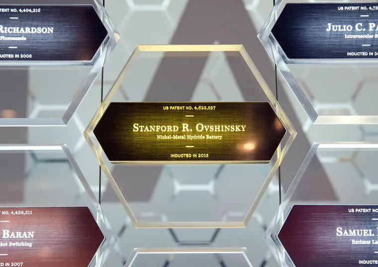 A hexagonal glass plaque displays Stanford R. Ovshinsky, Nickel-Metal Hydride Battery, Inducted in 2015 among other similar plaques featuring names and patents in a wall exhibit.