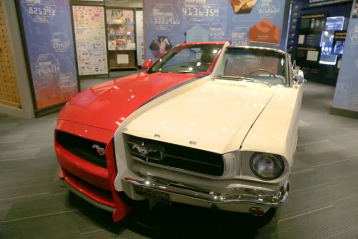 A car displayed in a museum is split down the middle, with the left half painted red and the right half painted white. The car is a classic Ford Mustang, and informational exhibits are visible in the background.