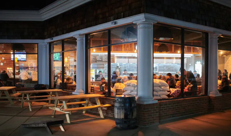 A restaurant with large windows shows people dining inside at night. Warm lights illuminate the interior, while empty picnic tables and a barrel are outside on the patio. Stacks of white bags are visible by the window.