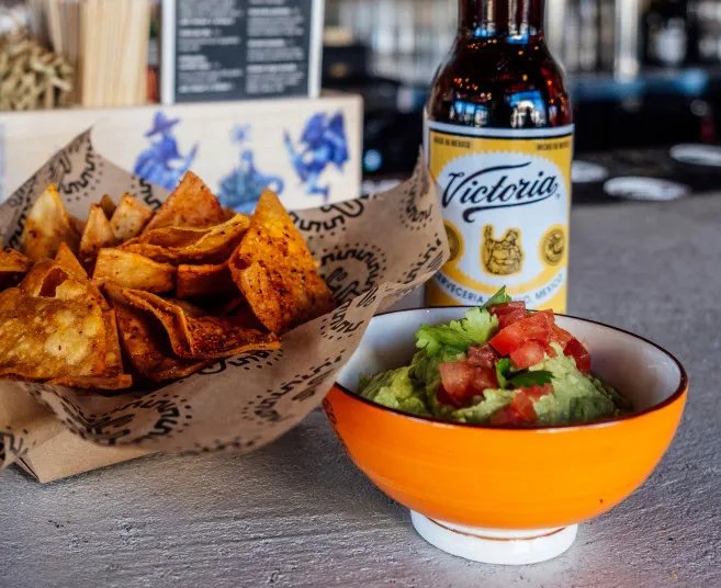 A bowl of guacamole topped with diced tomatoes sits next to a basket of tortilla chips and a bottle of Victoria beer on a restaurant table.