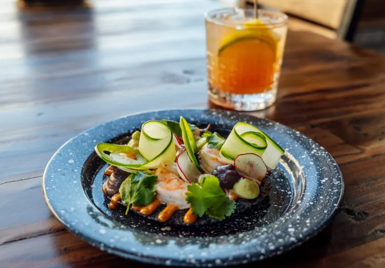 A dark plate holding a colorful dish with shrimp, cucumber ribbons, radish slices, cilantro, and sauce sits on a wooden table next to a glass of iced drink with a lemon or lime slice.