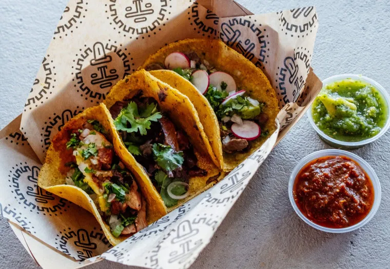 Three tacos filled with various meats, fresh cilantro, onions, and sliced radishes, served in a paper-lined tray with two small containers of green and red salsa on the side.