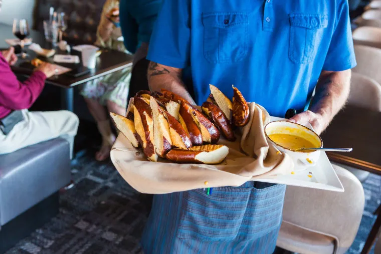 A server in a blue shirt holds a tray of sliced pretzels and a bowl of cheese dip in a restaurant, with diners seated at tables in the background.