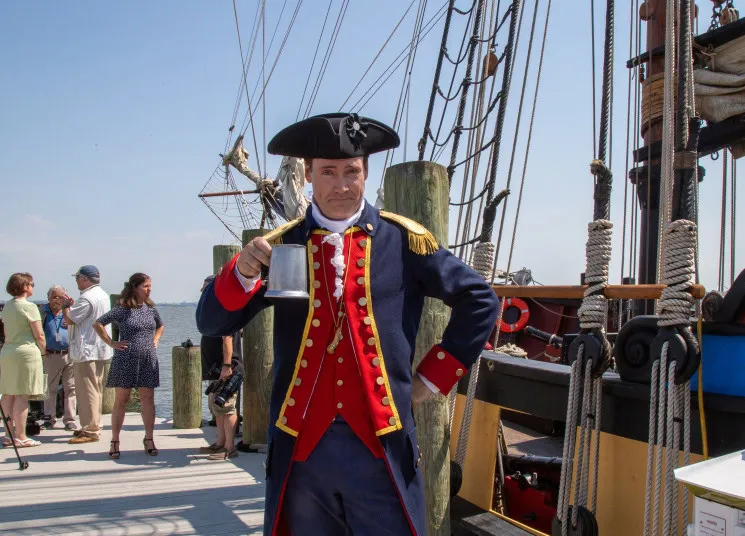A man in a historical military costume holds a metal mug and poses on a dock next to a tall ship, with several people and wooden posts in the background under a sunny sky.