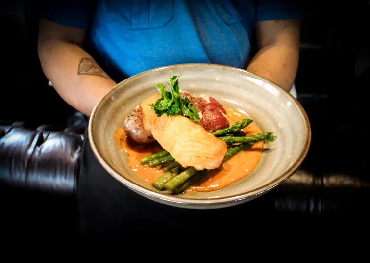 A person in a blue shirt holds a plate with a piece of cooked salmon, asparagus, red potatoes, and sauce, garnished with fresh herbs.