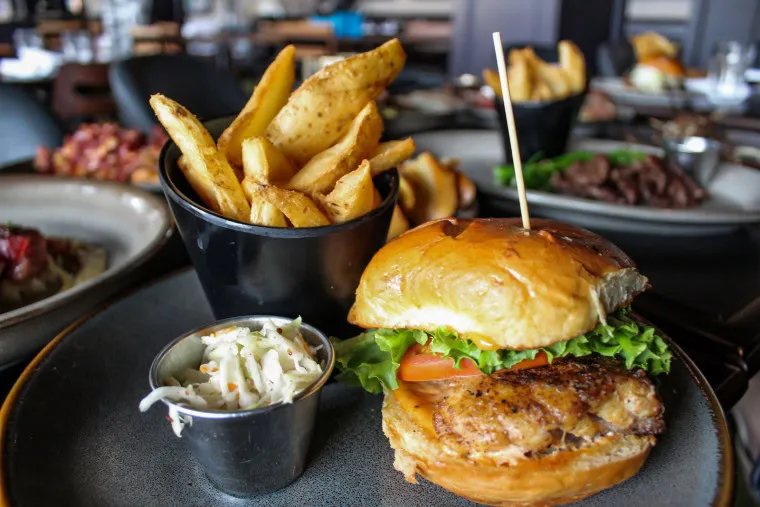 A plate with a grilled chicken sandwich featuring lettuce and tomato on a bun, served with a cup of coleslaw and a black bowl of thick-cut seasoned fries. Other food dishes are blurred in the background.