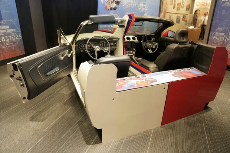 A car cut in half lengthwise is displayed indoors, showing the interior seats, dashboard, and steering wheel. Informational panels are attached to the sides, and the cars doors are open.