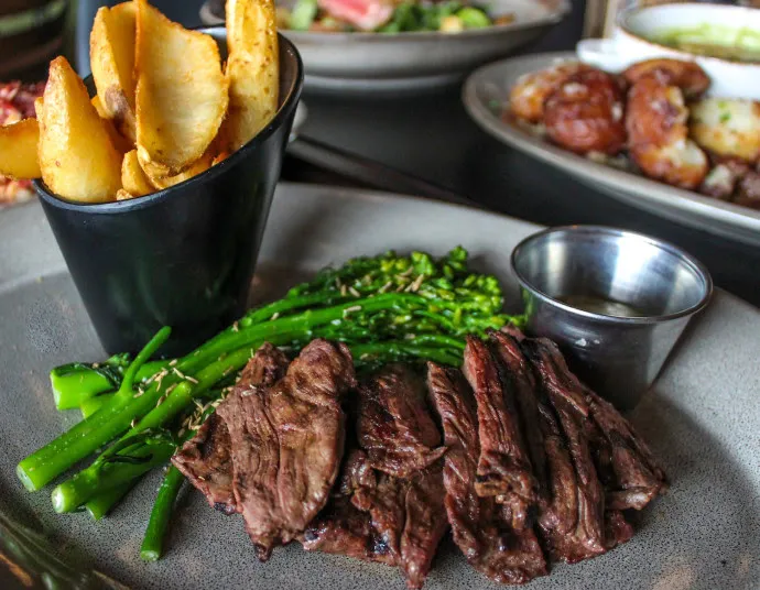 A plate with sliced grilled steak, broccolini, and a black cup filled with thick potato wedges. A small metal cup of sauce is on the side, with other dishes blurred in the background.