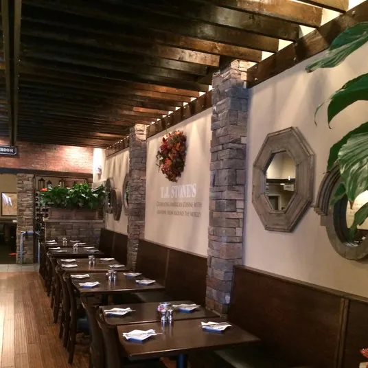 A cozy restaurant interior with wooden beams, stone columns, and a row of tables set with napkins and cutlery. Round mirrors and plants decorate the walls, and a wreath hangs above a sign reading T.J. STONES.