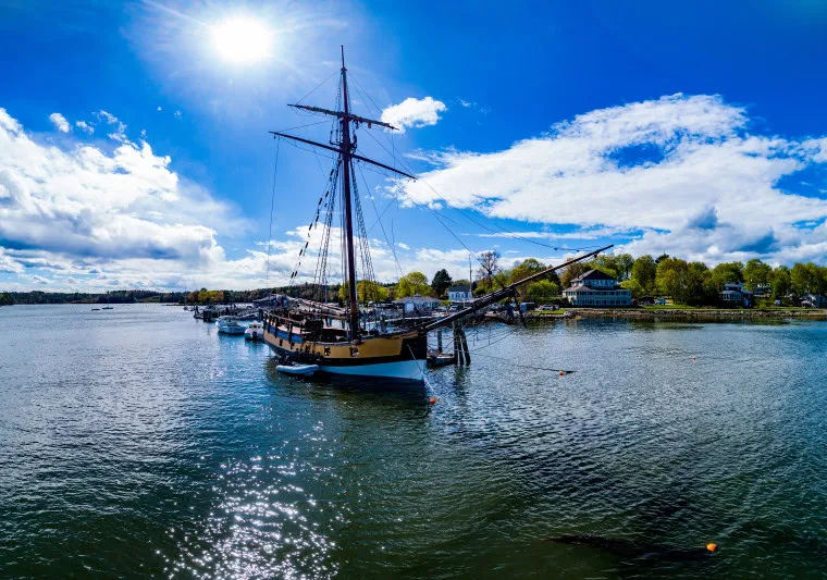 A historic sailing ship with tall masts is docked at a harbor on a sunny day with blue skies, scattered clouds, and surrounding waterfront buildings and trees. The sunlight reflects on the calm water.