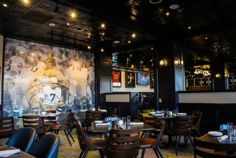 A modern restaurant with wooden tables and chairs, set for dining. A large mural of a football team decorates one wall, and sports memorabilia and photos are displayed throughout the space.