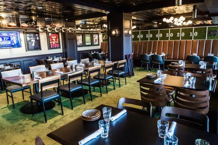 A sports-themed restaurant with green carpet, tables set with glasses and napkins, sports memorabilia and TVs on the walls, and booth seating under a scoreboard-style backdrop.