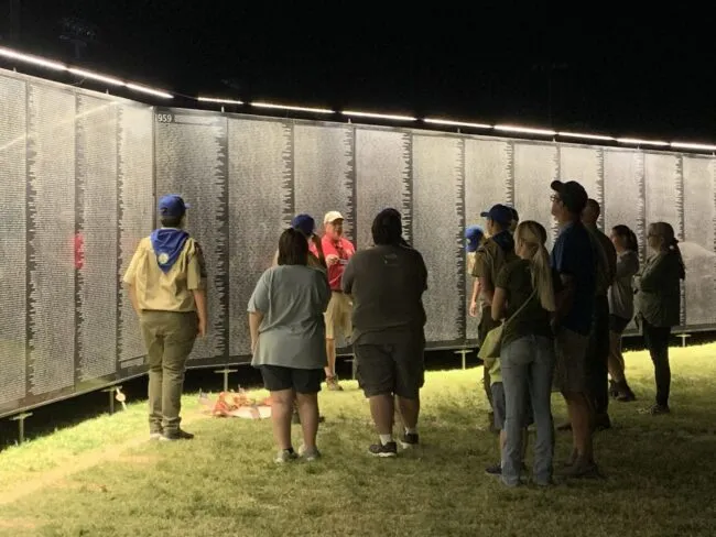 A group of people, including scouts, stand in front of a large illuminated wall displaying many engraved names at night, observing and discussing the memorial.
