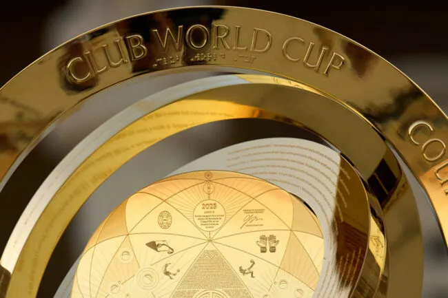 A close-up of the gold FIFA Club World Cup trophy, showing the engraved text CLUB WORLD CUP and intricate designs with detailed patterns and inscriptions.