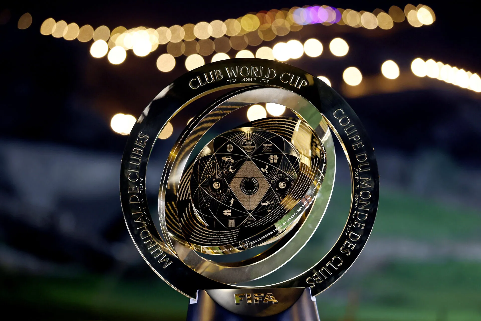 A close-up of the FIFA Club World Cup trophy, featuring a circular design with engraved text, set against a blurred background of lights at night.
