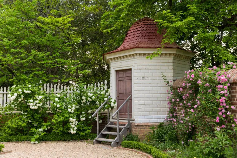 A small white brick garden shed with a red shingled roof and wooden steps sits among lush green trees, blooming bushes, and a white picket fence lined with white and pink flowers.