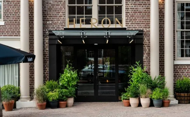 Entrance to a building with “HERON” sign above glass doors, surrounded by potted green plants and flanked by brick columns. The address number 699 is visible to the left of the doors.
