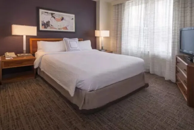 A hotel room with a neatly made king bed, two wooden nightstands with lamps, a framed artwork on a dark accent wall, a window with sheer curtains, and a TV on a wooden dresser.