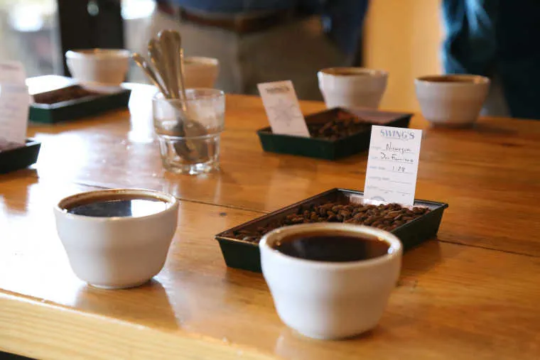 Several cups of brewed coffee and trays of coffee beans with labels are arranged on a wooden table, with spoons in a glass nearby, suggesting a coffee tasting or cupping session.