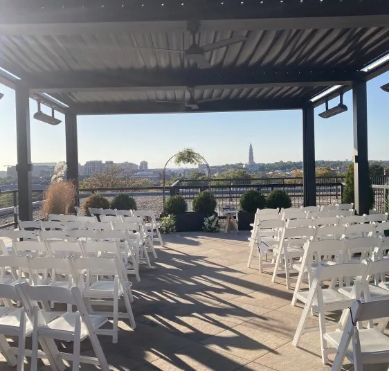 Rows of white chairs are arranged under a pergola on a rooftop patio, facing a circular wedding arch with greenery, overlooking a cityscape in the background under a clear sky.