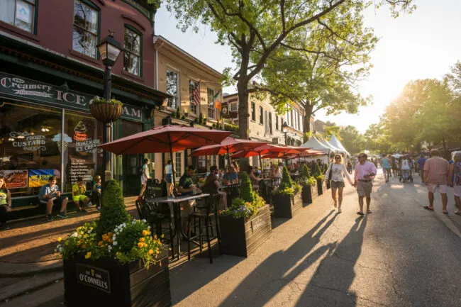 People enjoy outdoor dining under red umbrellas on a lively street lined with restaurants, including an ice cream shop, on a sunny day. Trees and flower planters decorate the scene as pedestrians walk by.
