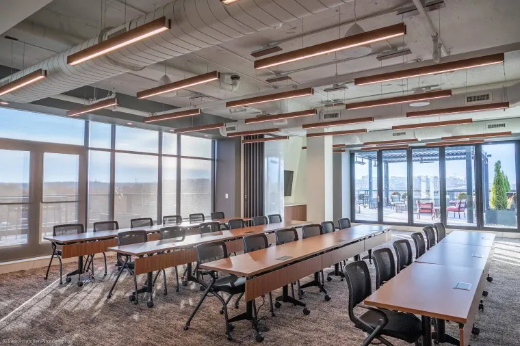A modern conference room with large windows, long wooden tables, black chairs on wheels, and contemporary ceiling lights. The room opens to an outdoor patio with city views in the background.
