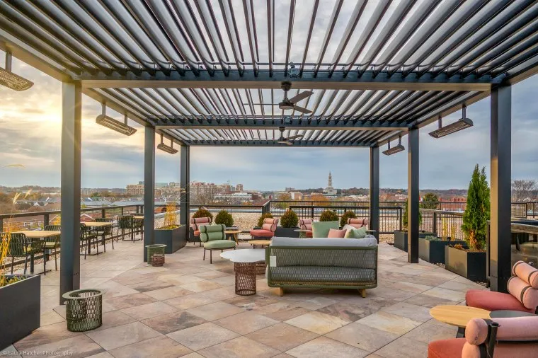A rooftop terrace with modern pergola, outdoor sofas, chairs, and tables overlooks a cityscape at sunset. Potted plants decorate the space, and warm sunlight filters through the pergola slats.