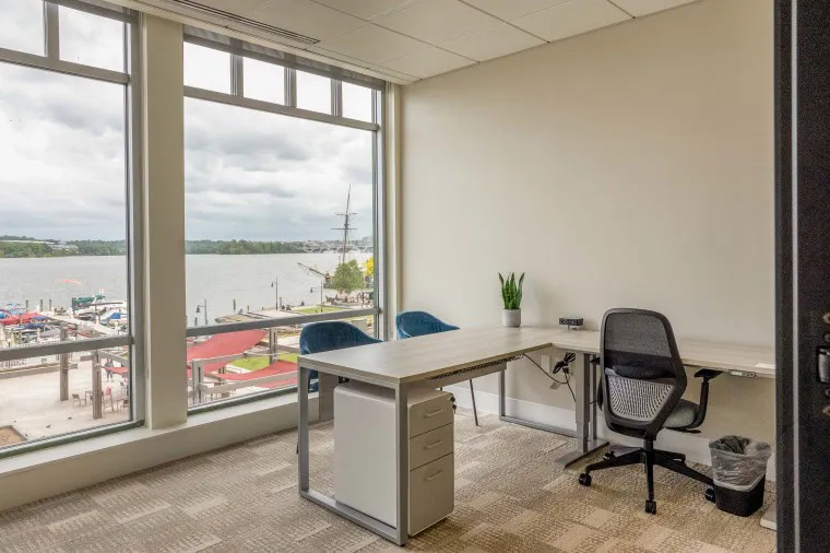 A modern office with a desk, office chair, two blue chairs, a potted plant, and large windows overlooking a waterfront with boats and a cloudy sky.
