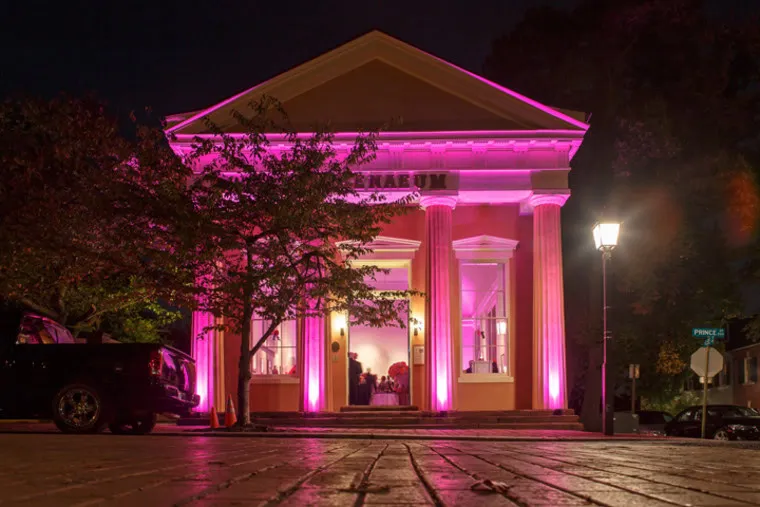A historic building with tall columns is illuminated by pink lights at night. Trees and a streetlamp are visible nearby, along with a car parked in front and a street sign on the corner.
