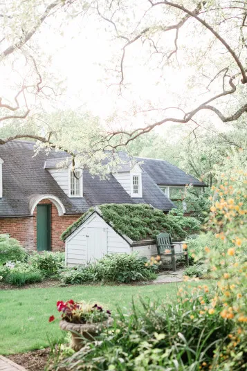 A charming house with brick and white siding sits among lush gardens. A green door and roof contrast with blooming flowers, leafy trees, and a green chair, creating a peaceful, sunlit garden scene.