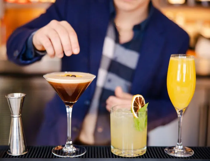 A bartender in a blue blazer prepares cocktails, including an espresso martini, a citrus drink garnished with mint and dried lime, and a mimosa, lined up on the bar.