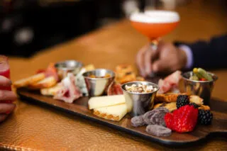 A wooden board with assorted cheeses, cold cuts, crackers, berries, chocolates, and small dishes, with two people holding colorful cocktails in the background.