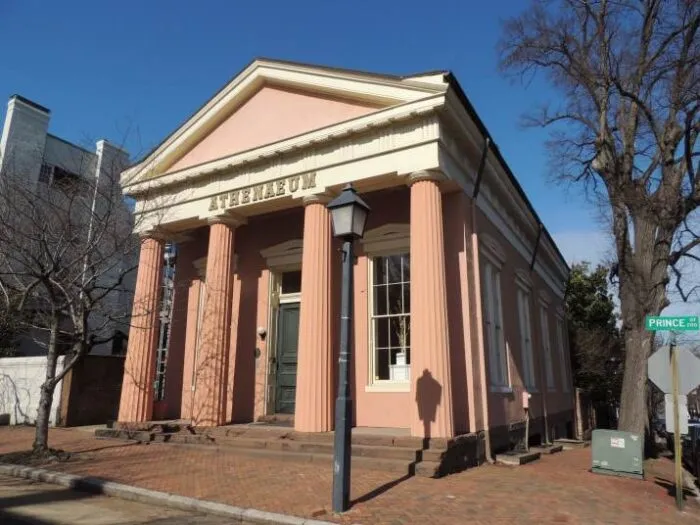 A historic pink and white Greek Revival building with columns and the word ATHENAEUM above the entrance, located on a brick sidewalk at the corner of Prince Avenue and another street.