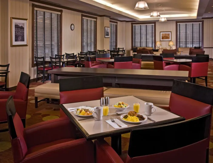 A dining area with red and black chairs and tables, set with plates of food, mugs, and utensils. The room is well-lit, has wooden blinds on the windows, and several empty tables and chairs in the background.