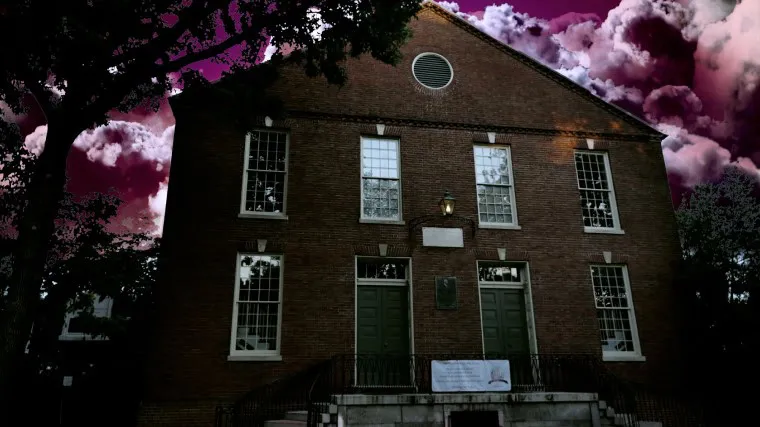 A dark brick building with tall windows and double green doors stands against a dramatic sky filled with purple and pink clouds, creating a mysterious and moody atmosphere. Trees partially frame the scene.