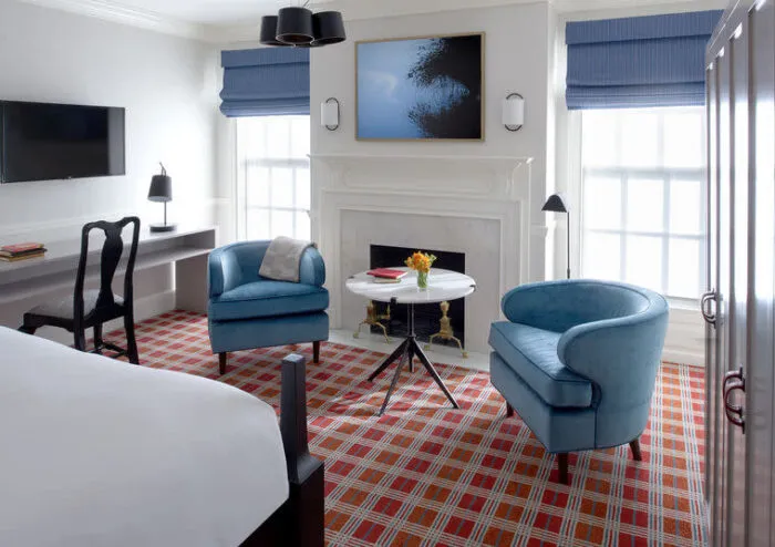 A bright hotel room with two blue armchairs around a small white table, a fireplace, a desk with a chair, a flat-screen TV, and large windows with blue blinds. The carpet is red and checkered.
