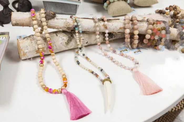 Four beaded necklaces with colorful tassels and beads are displayed draped over birch logs on a white surface. The necklaces feature various bead colors, including natural, pink, and orange tones.