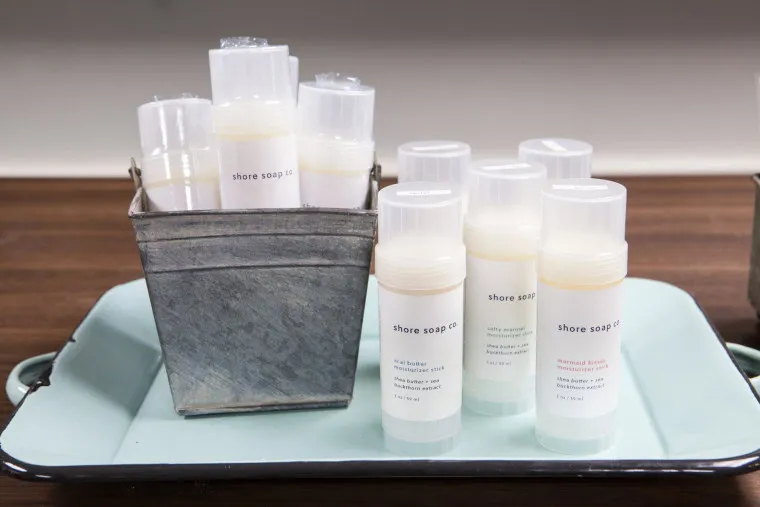 Several tubes of Shore Soap Co. lip balm are displayed on a light blue tray, with some upright in a small metal container and others standing on the tray, all against a wooden surface background.