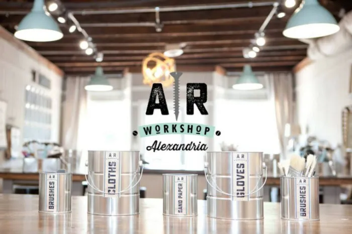 Bright workshop interior with metal cans labeled “BRUSHES,” “CLOTHES,” “SAND PAPER,” and “GLOVES” on a wooden table. Centered overlay text reads “AR Workshop Alexandria.” Exposed beams and lights above.