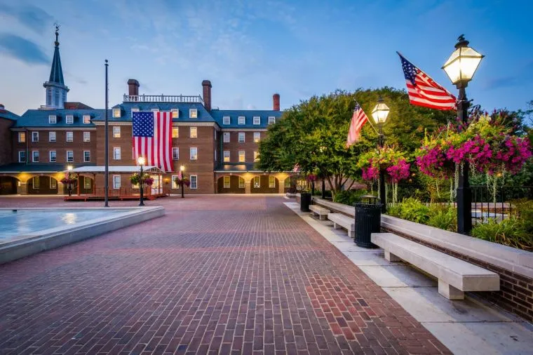 A brick-paved plaza with American flags, lamp posts, benches, hanging flower baskets, and a large brick building with a steeple in the background at dusk.