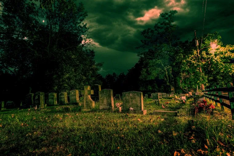 A dark, eerie cemetery at night under cloudy skies. Old tombstones are scattered across overgrown grass, with trees and a glowing streetlamp casting dramatic shadows.