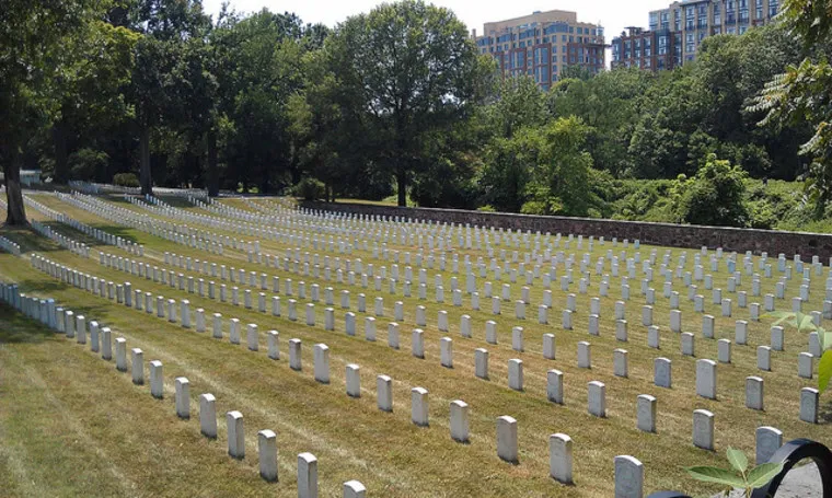 Rows of white headstones line a large, neatly kept cemetery with green grass, surrounded by trees and urban buildings visible in the background.