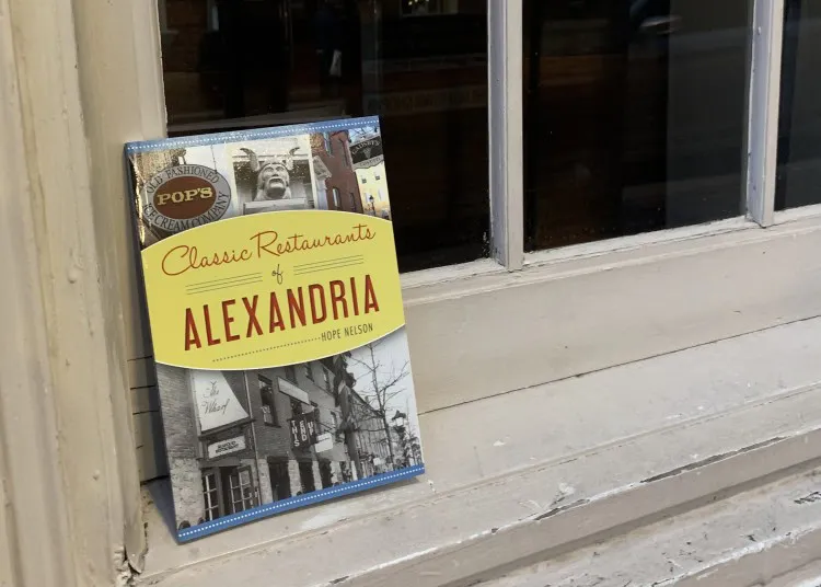 A book titled Classic Restaurants of Alexandria by Hope Nelson is propped against a beige wooden window sill. The cover features photos of restaurant signs and historic buildings.