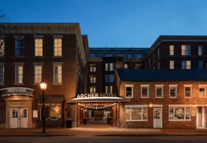 Archer Hotel exterior at dusk, with warm lights illuminating the entrance marquee; brick buildings surround the entrance, and two bicycles are parked outside on the sidewalk.